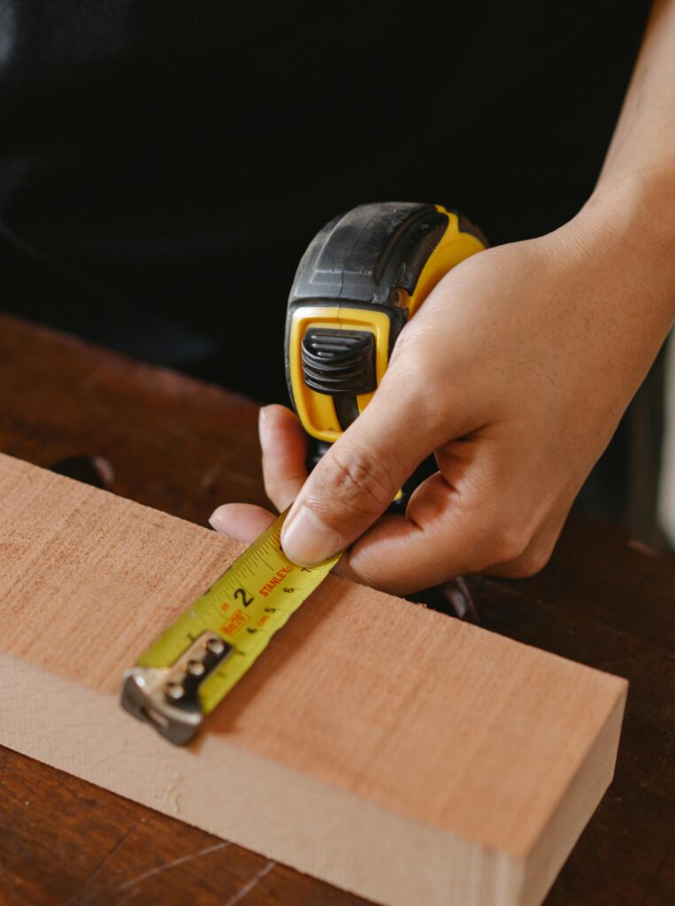 Close-up of a hand measuring wood with a tape measure in a workshop environment.